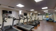 This fitness center is thoughtfully designed to provide a functional and comfortable exercise environment. The space features warm hardwood-style flooring that contrasts with the neutral-toned walls, creating an inviting atmosphere. The drop ceiling is equipped with recessed panel lighting that evenly illuminates the entire room, ensuring a bright and energizing workout space.

The cardio equipment lineup is a standout feature, with multiple treadmills and elliptical machines arranged in a row to maximize the use of floor space. Each machine is fitted with individual display consoles for tracking workout metrics. Wall-mounted flat-screen televisions are positioned at strategic points, allowing exercisers to stay entertained during their sessions.

A mirrored wall section at the far end of the room visually expands the space and allows users to monitor their form. A glass partition is also visible, potentially separating additional workout or utility areas. The overall layout is clean and organized, with sufficient spacing between machines to ensure user comfort and safety, making this fitness center a practical and well-appointed facility.