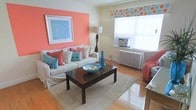 This single-floor space makes a bold first impression with its lively and thoughtfully coordinated interior design. The living area serves as the heart of the space, anchored by a striking coral accent wall that adds warmth and personality to the room. A comfortable white slipcovered sofa is dressed with an eye-catching mix of coral and turquoise throw pillows, reinforcing the space's cohesive color story. A dark wood coffee table with drawer storage sits atop a plush neutral area rug, providing both function and contrast against the warm hardwood flooring that runs throughout. Natural light flows in through a window dressed with a playful polka-dot valance in matching coral and turquoise tones, complementing the overall aesthetic. A classic white radiator beneath the window adds a charming, vintage character to the room, while a window air conditioning unit ensures year-round comfort. A tall sculptural floor lamp provides ambient lighting, and a framed blue abstract artwork on the coral wall ties the color palette together elegantly. Decorative accents, including a blue glass vase and a bowl centerpiece, add finishing touches that make the space feel curated and inviting. The single-floor layout offers a practical and comfortable living environment with a distinct, cheerful character.