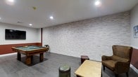 This single-floor recreational space is thoughtfully designed to provide a comfortable and engaging environment for leisure and entertainment. The room's most striking architectural feature is a full-length textured stone accent wall finished in light grey tones, lending the space a modern yet rustic character. Recessed ceiling lighting is evenly distributed throughout, casting a clean, well-lit ambiance across the entire area. The flooring is a durable dark grey wood-look material that complements the neutral palette of the walls and ceiling. A full-size pool table with a green felt surface serves as the centerpiece of the room, making it an ideal gathering spot for recreational activity. The space is further furnished with casual seating, including a cushioned chair and a low wooden table, encouraging a relaxed lounge atmosphere. Industrial-style accent pieces, such as a cylindrical metal stool and a compact trunk-style side table, add an eclectic, urban edge to the décor. Dark wood trim details along the upper walls provide a warm contrast to the lighter stone surfaces. Overall, the space balances a contemporary aesthetic with functional recreational amenities.