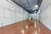 Located at 752 S Main Street, Unit 113, in the heart of downtown Los Angeles, this single-floor commercial unit offers a versatile, open-plan layout well-suited for a variety of uses, including retail, studio, or gallery space. The unit's industrial-modern aesthetic is defined by its exposed concrete ceilings with visible structural beams, ductwork, and conduit, all painted in a dark tone that contrasts sharply with the smooth, light gray walls throughout the main area. Polished hardwood floors run the length of the expansive open floor plan, reflecting the pendant lighting suspended from the ceiling above and lending a warm, refined quality to the otherwise raw industrial character of the space. Numerous electrical conduits and outlets are distributed along the walls, offering practical flexibility for a range of build-out configurations. Natural light enters through a large storefront-style entrance at the front of the unit, providing street visibility and a welcoming entry. Toward the rear, a partitioned section creates a more enclosed auxiliary area, while a private restroom with tile flooring and green accent walls adds functional convenience. The combination of open volume, durable finishes, and ample electrical infrastructure makes this unit a highly adaptable space in a prime downtown Los Angeles location.