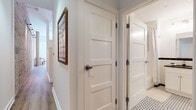 Located at 15 New St in West Hartford, Connecticut, this single-floor residence offers a thoughtfully designed open layout with warm, contemporary finishes throughout. The kitchen is a standout feature, showcasing rich honey-toned cabinetry, dark granite countertops, and a full suite of stainless steel appliances including a range, microwave, dishwasher, and French door refrigerator. A breakfast bar with a granite overhang and industrial-style metal stools provides casual seating, while an exposed brick accent wall adds a striking urban character. Light wood-style flooring flows seamlessly through the kitchen and into the main living areas, complemented by a ceiling fan overhead. The living area is bathed in natural light through large, black-framed industrial windows that frame views of the surrounding West Hartford streetscape, creating an airy and inviting atmosphere. The bathroom presents a timeless aesthetic with classic black-and-white penny tile flooring, a contrasting dark tile border, a white vanity with a dark countertop, and a shower-tub combination. The hallway features clean Shaker-style panel doors and warm wood flooring, with a framed art piece adding a personal touch. The overall ambiance blends industrial loft character with comfortable residential warmth.
