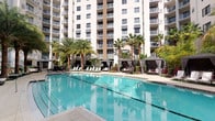 This outdoor amenity area showcases a generously sized swimming pool with crystal-clear turquoise water, designed to evoke a luxurious resort atmosphere. The pool deck is thoughtfully appointed with rows of lounge chairs ideal for sunbathing, while a series of elegant private cabanas with flowing curtains offer shaded retreats for relaxation and privacy. Towering palm trees are strategically placed throughout the space, providing a lush tropical ambiance and natural shade. Vibrant tropical plantings and well-maintained landscaping add color and life to the surroundings, enhancing the overall resort-like feel. The pool area is framed by a striking multi-story building, whose numerous balconies overlook the amenity space, offering elevated views of the pool and grounds below. The open-air design allows for abundant natural light throughout the day, creating a bright and inviting environment. The combination of thoughtful landscaping, stylish furnishings, and a spacious pool makes this outdoor space a standout feature, perfectly suited for leisure, social gatherings, and relaxation in a warm, sun-drenched setting.
