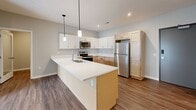 This single-floor unit presents a clean, modern aesthetic defined by neutral grey walls, wood-look vinyl plank flooring, and an open layout that promotes a sense of spaciousness. The kitchen serves as a central hub, anchored by a generous island with a crisp white countertop, an undermount double sink, and a stainless steel dishwasher. Light maple cabinetry contrasts with white upper cabinets, while stainless steel appliances—including a refrigerator, range, and microwave—add a polished finish. Two pendant lights hang above the island, complementing recessed lighting throughout. The kitchen opens directly into the living area, which features vertical blinds covering a sliding door that allows natural light to flow in. The bedrooms are well-proportioned, each with wood-look flooring, ceiling fans, and ample natural light from standard windows with blinds. The bathroom is neatly appointed with a light maple vanity, white countertop, a walk-in shower with built-in shelving, and wood-look flooring. A laundry area is also present within the unit, as indicated by the floor plan. Overall, the space offers a functional, contemporary design with cohesive finishes throughout.