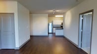 Located at Flats At 146, this single-floor unit offers a thoughtfully designed open-concept layout that seamlessly connects the living area to the kitchen. The living space is bright and inviting, featuring rich dark hardwood-style flooring that contrasts beautifully with the soft, neutral-toned walls, creating a clean and modern aesthetic throughout.

The kitchen is efficiently designed with white cabinetry that provides ample storage, a stainless steel refrigerator, a built-in microwave, and dark countertops with a sink positioned along the counter peninsula. The peninsula-style layout creates a natural division between the kitchen and living area while maintaining an open, airy feel. Overhead, a fluorescent light fixture ensures the kitchen is well-lit for cooking and meal preparation.

Track lighting in the main living area adds a contemporary touch and offers directional illumination to suit various needs. The smooth, neutral-painted walls throughout the unit provide a versatile backdrop, making it easy to personalize the space. The overall ambiance is modern, clean, and functional, ideal for those seeking a comfortable and low-maintenance living environment at Flats At 146.