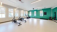 Located in North Charleston, South Carolina, this single-floor fitness facility offers a well-appointed workout environment divided into distinct functional zones. The cardio area is characterized by a bold teal accent wall flanked by multiple windows fitted with roller shades, allowing controlled natural light to fill the space. Ceiling fans and linear LED lighting ensure consistent illumination and comfort throughout. The smooth, light-colored resilient flooring provides a clean, durable surface suitable for a range of exercises. Equipment in this zone includes stationary cycling bikes with digital displays and rowing machines. An adjoining studio area features a full-length mirrored wall, which enhances the sense of openness and supports movement-based workouts. This section is distinguished by a richly patterned blue and white accent wall, a built-in white shelving unit framed in navy, and organized storage for stability balls, hand weights, and accessories. Ceiling fans continue into this zone, maintaining a consistent design language. Together, the two areas create a cohesive, modern fitness environment that balances energetic design elements with functional, practical amenities suited for diverse workout routines.