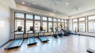 Located at 255 Williamsburg Pkwy in Fate, TX, this fitness center offers a comprehensive workout environment designed to support a wide range of exercise routines. The single-floor space is thoughtfully laid out to maximize functionality and comfort for users of all fitness levels.

The facility is bathed in natural light, courtesy of an expansive bank of large, multi-pane windows that line the perimeter walls and provide views of the surrounding community. Elevated ceilings with large ceiling fans enhance air circulation and contribute to an open, airy atmosphere. Recessed lighting throughout ensures the space is well-lit at all hours.

The cardio zone features multiple treadmills, stationary bikes, stair climbers, and elliptical machines, all positioned to face the windows for an engaging workout experience. The strength training area is equally well-appointed, offering a multi-station cable machine, a full rack of dumbbells, weight plates, and benches for free weight exercises. A wall of mirrors in the strength zone supports proper form and visually expands the space.

Additional amenities include built-in storage cabinetry, organized accessory storage for items such as resistance balls and exercise mats, and a wall-mounted television screen. Light wood-tone flooring runs throughout, providing a clean, modern aesthetic. A glass-paneled entry door connects the fitness center to adjacent community spaces.