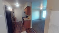 This single-floor space is designed for practical, comfortable living. The kitchen is outfitted with warm-toned wooden cabinetry, both upper and lower, providing ample storage. A light-colored countertop runs along the workspace, complemented by a black range and a refrigerator. Dark hardwood-style flooring runs throughout the kitchen area, adding a rich, grounded aesthetic. A range hood is mounted above the stove, enhancing the kitchen's functionality. The kitchen flows openly into the adjacent living area, which transitions to plush, dark carpeting. The living space benefits from a window that introduces natural light, brightening the room and creating an inviting atmosphere. A ceiling fan is centrally mounted, offering both air circulation and supplemental lighting. The textured ceilings are consistent throughout, and neutral wall tones provide a versatile backdrop. A hallway visible from the kitchen suggests additional rooms beyond the main living areas. Overall, the space strikes a balance between functional design and comfortable everyday living.