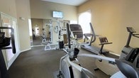 This fitness center offers a comprehensive workout environment designed to support a wide range of exercise routines. The space is outfitted with a solid selection of equipment, including elliptical machines, treadmills, and a multi-station cable weight machine, providing options for both cardiovascular and strength training workouts. A wall-mounted flat-screen television is positioned prominently at the front of the room, offering entertainment during exercise sessions. Large mirrors line the walls, enhancing the sense of space and allowing users to monitor their form throughout their workouts. Natural light filters in through a window, brightening the room and creating an inviting atmosphere. The flooring features durable, low-pile carpet suited for a fitness environment. Neutral, warm-toned walls contribute to a clean and focused ambiance. An emergency exit sign is visible, reflecting attention to safety throughout the space. Overall, this fitness center is thoughtfully arranged to maximize functionality while maintaining an open, comfortable feel that encourages regular use.
