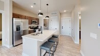 This well-appointed single-floor residence showcases a thoughtful, modern design with a neutral color palette and light wood-look flooring that flows seamlessly throughout. The open-concept kitchen and living area forms the heart of the home, centered around a generous white quartz island with an undermount sink, matte black fixtures, and seating for multiple guests. Rich dark-walnut shaker-style cabinetry lines the kitchen walls, complemented by a full suite of stainless steel appliances, including a French door refrigerator, range, and built-in microwave. Stylish pendant lights hang above the island, adding both function and visual warmth. The adjacent living area benefits from a bank of large windows that invite abundant natural light and an open, airy feel. Two well-proportioned bedrooms feature neutral toned walls, large windows with blinds, and easy access to the bathrooms. The primary bathroom offers a dark wood vanity with white quartz countertop and a tub-shower combination, while the secondary bathroom mirrors the same refined aesthetic. A dedicated in-unit laundry area with a full-size washer and dryer adds everyday convenience. Overall, the space presents a clean, contemporary ambiance ideal for modern living.