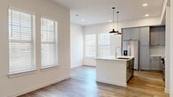 This well-appointed single-floor residence offers a thoughtfully designed layout that maximizes comfort and functionality. The open-concept kitchen serves as the heart of the home, showcasing sleek gray shaker-style cabinetry extending to the ceiling, a white quartz waterfall island with an integrated sink, stainless steel appliances including a French door refrigerator, range, built-in microwave, and a wine cooler. A white penny-tile backsplash and stylish black pendant lights add refined character, while multiple windows flood the space with natural light. Light wood-tone luxury vinyl plank flooring flows throughout the main living areas, creating a seamless and cohesive aesthetic. Hallways connect the living spaces efficiently, with clean white walls and paneled doors featuring matte black hardware. The spacious bedroom features plush carpeting, dual windows with blinds for natural light and privacy, and a ceiling fan fixture. A dedicated closet provides practical storage. The bathroom is finished with large-format tile flooring, a glass-enclosed walk-in shower with subway tile surround, a gray vanity with a white countertop, a lighted mirror, and matching cabinetry. The overall ambiance is bright, clean, and modern throughout.