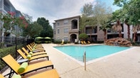 This single-floor residence is part of a multi-story building complex, offering both thoughtfully designed interior spaces and access to impressive outdoor amenities. The community's outdoor area centers around a large, sparkling swimming pool framed by an expansive sun deck lined with yellow and green lounge chairs and shaded umbrella seating, all set against a backdrop of mature trees and lush landscaping.

Inside, the unit presents an eclectic yet refined aesthetic. The interiors feature polished concrete flooring throughout, lending an industrial-chic character that is balanced by warm, neutral wall tones. A standout element is the dining area, anchored by a striking round green marble pedestal table accompanied by woven rattan chairs, all set atop a natural fiber area rug. A large framed abstract artwork in soft earth tones adorns the wall, adding an artistic focal point to the space.

The unit also incorporates distinctive architectural details, including a sliding barn door on an exposed black iron rail system, which adds both functional versatility and visual interest. Vintage-style double doors with distressed patina lead into an adjacent room, where additional framed artwork and a teal accent wall hint at further creative interior choices. Recessed lighting throughout ensures a clean, uncluttered ceiling plane, while the overall design balances artistic personality with understated sophistication.