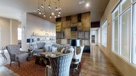 This single-floor space showcases a thoughtfully curated interior that blends rustic character with contemporary sophistication. The centerpiece of the room is a dramatic floor-to-ceiling accent wall constructed from reclaimed wood panels and aged materials arranged in a patchwork pattern, creating a bold focal point that anchors the entire space.

The seating area is elegantly appointed with tufted gray upholstered sofas and wingback chairs, arranged around a glass-topped coffee table and complemented by a striking black and silver table lamp. A rich, patterned area rug in deep red tones adds warmth and definition to the lounge area.

Overhead, a cascading pendant light fixture featuring exposed Edison-style bulbs adds an industrial yet artistic touch, drawing the eye upward to the generously proportioned ceilings. Along one wall, built-in shelving displays carefully arranged decorative accessories, adding a curated, boutique-like quality to the space.

Large windows along the perimeter flood the interior with natural light, enhancing the warm tones of the hardwood flooring that runs throughout. The overall ambiance is inviting and stylish, balancing raw, textural elements with refined furnishings.