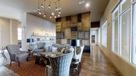 This single-floor space showcases a thoughtfully curated interior that blends rustic character with contemporary sophistication. The centerpiece of the room is a dramatic floor-to-ceiling accent wall constructed from reclaimed wood panels and aged materials arranged in a patchwork pattern, creating a bold focal point that anchors the entire space.

The seating area is elegantly appointed with tufted gray upholstered sofas and wingback chairs, arranged around a glass-topped coffee table and complemented by a striking black and silver table lamp. A rich, patterned area rug in deep red tones adds warmth and definition to the lounge area.

Overhead, a cascading pendant light fixture featuring exposed Edison-style bulbs adds an industrial yet artistic touch, drawing the eye upward to the generously proportioned ceilings. Along one wall, built-in shelving displays carefully arranged decorative accessories, adding a curated, boutique-like quality to the space.

Large windows along the perimeter flood the interior with natural light, enhancing the warm tones of the hardwood flooring that runs throughout. The overall ambiance is inviting and stylish, balancing raw, textural elements with refined furnishings.