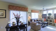 This single-story space presents an open-concept floor plan that integrates the dining and living areas into one cohesive, well-appointed environment. The dining area features a dark-toned table set with decorative place settings and accented by a bold floral arrangement, adding a vibrant touch to the space. Large vertical blinds on sliding glass doors allow abundant natural light to flood the interior, while decorative valances in warm terracotta tones with a leaf motif provide a consistent design thread throughout the space.

The living area is furnished with a leather sofa and complementary wicker-style seating, creating a comfortable and visually layered arrangement. A ceiling fan with a light fixture overhead enhances airflow and ambient lighting. Additional windows with matching valances continue to brighten the room naturally. Soft, neutral-toned carpeting runs throughout the living space, contributing to its warm and relaxed ambiance. Decorative wall art, floor lamps, and coordinated accessories complete the interior, reflecting a cohesive and thoughtfully styled aesthetic suitable for both everyday living and entertaining.
