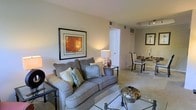 This single-story space in Brandywine presents an open-concept layout that seamlessly connects the living and dining areas, creating a cohesive and functional flow throughout. The living area is anchored by a plush, generously sized sofa adorned with layered throw pillows in soft blue and green tones, complemented by sleek glass-topped side and coffee tables that add a contemporary touch. A framed artwork with warm amber tones decorates the wall, contributing to the inviting atmosphere enhanced by a classic table lamp that casts soft, ambient light.

The light-toned flooring — which appears to be a polished tile or similar smooth surface — extends throughout both areas, reinforcing a sense of continuity and easy maintenance. The dining area, visible from the living space, features a set of elegant chairs surrounding a dining table, with additional framed art and candle accents that add refinement.

The neutral color palette of warm beige and cream tones on the walls and flooring creates a calm, versatile backdrop. Recessed ceiling details and visible HVAC vents suggest thoughtful interior planning. Overall, this space delivers a balanced combination of comfort, functionality, and understated elegance.