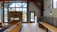 Located at 1505 E Burnsville Pkwy in Burnsville, Minnesota, this single-floor clubhouse combines contemporary design with warm, natural materials to create an inviting communal gathering space. The interior is defined by dramatic vaulted ceilings supported by exposed timber beam trusses, lending the space a rustic yet refined architectural character. Light hardwood flooring runs throughout, complementing the neutral gray walls and natural wood accents.

The main gathering area features a full-height stone fireplace flanked by floor-to-ceiling black-framed windows that flood the interior with abundant natural light and offer open views of the surrounding greenery. Comfortable seating, including a leather sofa and upholstered lounge chairs alongside natural wood stump side tables, creates a relaxed and welcoming atmosphere.

Built-in banquette seating with a wood-topped dining table provides a dedicated area for casual meals or meetings. A kitchenette area is equipped with gray cabinetry, a farmhouse sink, white subway tile backsplash, pendant lighting, and a wall-mounted television. Multiple black-framed glass doors provide seamless access to an outdoor patio, extending the usable space outdoors. The overall design balances modern finishes with organic textures, resulting in a functional and aesthetically cohesive community amenity space.