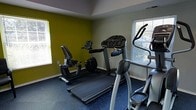 This single-floor fitness room is purpose-built for cardiovascular training and general exercise. The space is outfitted with a dedicated selection of cardio equipment, including a treadmill, an elliptical trainer, and a recumbent stationary bike — providing users with a variety of low-impact and high-intensity workout options. The room features durable blue rubber flooring, ideal for athletic use and easy maintenance. The walls are painted in a two-tone scheme, combining a bold chartreuse-green accent wall with neutral gray surroundings, lending the space an energetic and motivating atmosphere. Natural light filters in through multiple windows fitted with horizontal blinds, brightening the room and creating a pleasant environment for exercise. The layout is efficient and well-organized, allowing for comfortable movement between machines. The ceiling features a subtle tray detail, adding a modest architectural element to the otherwise utilitarian space. Overall, this fitness room offers a functional and focused environment for residents or users looking to maintain an active routine without leaving the building.