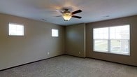 This two-story space offers a functional and straightforward layout with clean, neutral finishes throughout. The pictured room exemplifies the interior's understated design, featuring wall-to-wall carpeting in a light beige tone that adds warmth underfoot. The walls are finished in a muted olive-beige hue, complemented by dark baseboards that provide a subtle contrast. A ceiling fan with dark wood-toned blades and an integrated light fixture is centrally mounted, serving both ventilation and ambient lighting needs. Two windows bring in natural light — a smaller square window on one wall and a larger window on the adjacent wall — offering views of what appears to be a green outdoor landscape. Electrical outlets are visible along the lower portion of the walls, and a ceiling vent indicates a climate control system is in place. The room's proportions feel open and versatile, suitable for a variety of uses. With two floors in total, the space provides separation between living areas, allowing for flexible use of each level. The overall ambiance is calm and neutral, offering a blank canvas well-suited for personalization.