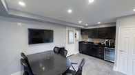 This single-floor space offers a well-designed, open layout that seamlessly combines a dining area and a kitchenette into one cohesive environment. The dining area is anchored by a dark granite-topped round table surrounded by matching black chairs, providing a comfortable and stylish setting. A large wall-mounted flat-screen television is prominently positioned on the neutral gray wall, making the space well-suited for both meetings and casual gatherings. Natural light filters in through a shuttered glass door, brightening the interior and adding a welcoming feel. The kitchenette is outfitted with rich, dark espresso cabinetry paired with dark countertops and a mosaic tile backsplash that adds a subtle decorative touch. A stainless steel sink and compact appliances complete the functional kitchen setup. Recessed lighting throughout the ceiling ensures even, modern illumination across the entire space. The light-toned tile flooring provides a clean contrast to the dark furnishings and cabinetry, contributing to a balanced and polished aesthetic. Overall, the space presents a contemporary, functional design ideal for a variety of uses.
