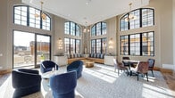 Located at 600 West State Street in Kennett Square, Pennsylvania, this stunning living room makes a bold architectural statement through its soaring double-height ceilings and expansive arched, grid-paned windows that flood the interior with abundant natural light. The generous open floor plan is thoughtfully arranged into distinct seating zones, offering both social and intimate gathering areas. A built-in striped bench sofa anchors the central window wall, complemented by plush, curved navy blue lounge chairs and sculptural brass accent tables that add a refined, contemporary flair. A dedicated dining nook features a round pedestal table surrounded by elegant upholstered chairs, set against a warm wood slat partition that adds texture and visual warmth. The entrance area is framed by a grand arched doorway with rich wood trim, reinforcing the space's upscale character. Globe pendant chandeliers and wall-mounted multi-globe sconces in brushed brass cast a warm, inviting glow. Soft neutral wall tones are offset by light hardwood flooring and plush area rugs. Large glass doors open to an adjacent outdoor terrace, seamlessly blending interior comfort with outdoor connectivity. The overall ambiance is polished, airy, and contemporary.