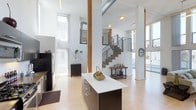 This striking two-floor space showcases a thoughtfully designed interior that blends industrial and contemporary elements. The main level is anchored by a generous open-plan living and kitchen area, where soaring double-height ceilings create an impressive sense of volume and openness. Warm wood-toned flooring runs throughout, complementing the exposed wooden structural columns that add architectural character and a natural warmth to the space. The kitchen features a sleek central island with a white countertop and dark cabinetry, ideal for both everyday use and entertaining. Exposed ductwork overhead nods to the building's industrial aesthetic, while large windows on multiple sides flood the interior with abundant natural light. A modern staircase with a metal railing connects the two floors, serving as both a functional element and a design focal point. The upper floor provides additional living space, maintaining the same clean, open aesthetic. Beyond the interior, a spacious rooftop terrace offers an expansive outdoor retreat under open skies, enclosed by sleek glass-panel railings. The terrace provides wide-open views of the surrounding neighborhood and adjacent buildings, making it a standout feature perfect for outdoor relaxation or hosting.