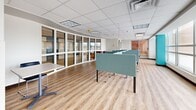 This contemporary open-plan workspace is designed to support a variety of working and studying styles within a well-organized, single-floor layout. The space features light-toned hardwood-style flooring that runs throughout, contributing to a clean and welcoming atmosphere. A drop tile ceiling with recessed and panel lighting ensures consistent, even illumination across the entire floor.

Individual workstations equipped with teal acoustic privacy dividers are arranged throughout the main area, providing focused work zones without fully enclosing the space. A lounge area with plush purple armchairs and a small table offers a comfortable setting for informal meetings or relaxed study. Large windows line the exterior wall, flooding the interior with natural light and providing outward views of the surrounding area.

Vibrant teal accent walls punctuate the otherwise neutral white-and-grey palette, adding a dynamic visual element. A wall-mounted flat-screen display is positioned at one end of the space, supporting presentations or shared media. Glass partition walls with white framing define a separate enclosed area at one end, maintaining visual openness while offering a degree of acoustic separation. Additional collaborative tables provide flexible workspace options. The overall design balances productivity and comfort, making it well-suited for co-working, study, or light office use.