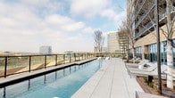 This expansive rooftop amenity deck offers a thoughtfully designed outdoor retreat elevated above the surrounding urban landscape. The centerpiece is a sleek, resort-style lap pool that stretches along the perimeter of the deck, bordered by large-format light grey pavers and equipped with contemporary white lounge chairs. Glass panel railings frame the pool area, preserving unobstructed panoramic views of the city skyline and open horizon beyond.

Adjacent to the pool, a pergola-covered outdoor kitchen provides a well-appointed space for entertaining, complete with a stainless steel grill, a granite-topped prep island with an integrated sink, and warm wood-slat overhead detailing. Raised planter beds clad in weathered Corten steel and warm brick add structured greenery throughout the deck, featuring ornamental grasses and seasonal plantings that soften the hardscape.

The building's exterior façade of light-toned brick and expansive floor-to-ceiling glass walls lines the deck, with wide French doors providing seamless indoor-outdoor access. Accessible pathways with metal railings connect various zones of the terrace, ensuring smooth circulation throughout. The overall ambiance is one of modern elegance, offering an open-air environment designed for relaxation, recreation, and social gatherings with striking skyline views as a constant backdrop.