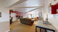 Located at 7520 W Amherst Ave in Denver, CO, this well-appointed single-floor community space offers a thoughtfully designed layout encompassing a living room, two dining areas, and a full kitchen. The living room features vaulted ceilings with exposed beams, recessed lighting, warm neutral carpeting, and a classic brick fireplace flanked by comfortable seating — creating an inviting communal atmosphere. Bold accent walls in deep red add visual character throughout the space. One dining area accommodates a large party with a substantial dark wood table and leather-upholstered chairs beneath skylight-style overhead lighting, while a second dining area offers a more intimate setting with a glass-top table and floor-to-ceiling windows that open to an outdoor deck, flooding the room with natural light and tree-lined views. The kitchen is efficiently outfitted with light wood cabinetry, dark laminate countertops, a double stainless steel sink, open wall-mounted shelving, and stainless steel appliances including a full-size refrigerator, all set on durable hardwood-style flooring. Throughout the space, crown molding, chair rail detailing, and abundant natural light contribute to a polished, welcoming ambiance suited for gatherings and events.