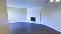 This single-story space presents a straightforward yet inviting layout centered around a prominent white-painted brick fireplace, which serves as the focal point of the main living area. The fireplace surround extends from floor to ceiling, adding architectural character and a sense of depth to the room. Dark hardwood flooring runs throughout, providing a rich contrast against the light-toned walls and white brick, and contributes to a grounded, warm atmosphere. The walls are finished in a soft neutral tone, lending the space a clean and versatile backdrop suitable for a variety of interior styles. A textured popcorn ceiling adds a classic residential quality overhead. The room is generously proportioned, offering ample open floor space that allows for flexible furniture arrangement. Subtle details such as standard electrical outlets along the baseboards and a ceiling fan suggest practical, functional living. The overall ambiance is simple and neutral — a blank canvas that balances comfort with understated design, making it well-suited for those looking to personalize the space to their own taste.