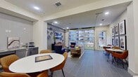 This well-appointed amenity lounge is a single-floor space designed to serve as a multifunctional gathering and recreation area. The floor is finished throughout in large-format slate-toned tiles, lending a cohesive and polished look to the entire space.

The lounge area is anchored by a wall-mounted flat-screen TV, complemented by bold blue sculptural wall art and a warm wood media credenza. Comfortable seating — including a plush gray sofa and vibrant navy blue accent chairs — creates an inviting space for relaxation and socializing. A standout architectural feature is the full-height glass panel garage-style door along one wall, which floods the lounge with natural light and offers clear views of an outdoor courtyard and pool area below.

Adjacent to the lounge is a casual dining zone with a round white table and tan leather chairs, perfectly positioned near a compact wet bar. The wet bar features gray cabinetry, a stainless steel sink, a dishwasher, and marble-look countertops — ideal for entertaining. A foosball table adds a recreational element nearby, while a cozy reading nook with an egg chair rounds out the space.

Recessed lighting and a stylish multi-arm chandelier provide warm, even illumination throughout, while curated artwork and decorative plants enhance the modern, welcoming ambiance of this amenity space.