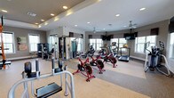 This fitness center is organized across a single level and thoughtfully divided into distinct workout zones to accommodate a wide range of training styles. The cardio area is outfitted with multiple treadmills, elliptical machines, and red cycling bikes, all positioned near large windows that allow abundant natural light to fill the space. Ceiling fans and ceiling-mounted flat-screen televisions enhance comfort during cardio sessions.

The strength training zone features a bold orange power rack with an extensive array of attachments, a dedicated bench press station, barbells with weighted plates, medicine balls, and a heavy punching bag — providing comprehensive options for resistance and functional training. A cable machine and additional weight equipment further expand training versatility. Large mirrors are strategically placed to support proper form and visually open the space.

The facility is finished with durable rubberized flooring throughout, recessed lighting, and tray ceiling details that lend a polished aesthetic. Colorful artwork accents the walls, adding visual interest. Entry to the fitness center is secured via a keypad access system, with sanitization supplies and waste disposal conveniently available at the entrance for hygiene maintenance.