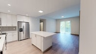 This well-designed single-floor residence offers a clean, contemporary aesthetic throughout, with light wood-tone flooring that flows seamlessly from room to room. The open-concept kitchen is a standout feature, equipped with a full suite of stainless steel appliances — including a French-door refrigerator, range, microwave, and dishwasher — complemented by crisp white cabinetry, a grey geometric tile backsplash, and a spacious central island with a white countertop, ideal for both meal preparation and casual dining. The kitchen opens directly into a generously sized living area, which is bathed in natural light from sliding glass doors that lead to an outdoor space with verdant views beyond.

The bedrooms are bright and comfortable, each featuring light wood flooring, large windows with blinds that invite natural light, and clean white walls that create a calm, neutral atmosphere. Additional rooms offer sliding door access to outdoor areas, further enhancing the connection to the exterior.

The bathrooms exhibit a polished, modern design, showcasing marble-look tile flooring, white quartz vanity tops, and elongated hexagonal wall tile in the tub and shower surrounds. A wood-framed mirror above a dual-sink vanity adds a warm accent to the otherwise crisp, all-white palette. Recessed lighting throughout the residence ensures consistent, even illumination in every space.