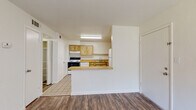 This single-floor unit offers a functional and well-organized layout with an open-concept design connecting the kitchen and living areas. The kitchen is equipped with light wood cabinetry, laminate countertops, a full suite of white appliances including an electric range, dishwasher, and refrigerator, and a tile floor. A peninsula counter separates the kitchen from the adjacent living and dining space, which features durable wood-look plank flooring throughout. The living area is bright and open, with a ceiling fan, a sliding glass door fitted with vertical blinds that allows for natural light, and a dedicated entryway. A doorway from the main living space leads to a walk-in pantry or storage closet, adding practical storage options. The bedroom is generously sized with soft carpeting, a ceiling fan with light fixture, a window with vertical blinds, and access to a walk-in closet with wire shelving. The bathroom includes a tiled floor, a full-size soaking tub and shower combination with chrome fixtures, and clean, neutral finishes throughout. The overall aesthetic is clean and neutral, with white walls and a simple, utilitarian design that provides a versatile canvas for personalization.