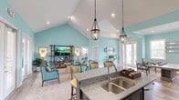 Located at 2821 Carolina Beach Rd in Wilmington, NC, this single-story clubhouse space exudes a welcoming coastal charm. The open-concept layout seamlessly connects a fully equipped kitchen island — featuring granite countertops, a double stainless steel sink, and bar seating with cheerful yellow accents — to a spacious lounge area ideal for gathering and relaxing. Soaring vaulted ceilings with crisp white finishes amplify the sense of openness, while the signature aqua-toned walls lend a refreshing, beach-inspired atmosphere throughout. Recessed lighting and elegant dark pendant lanterns hang from the peaked ceiling, providing warm, layered illumination. The lounge area is thoughtfully furnished with comfortable seating arrangements, including coastal-style upholstered chairs in coordinating teal tones, positioned around a flat-screen television mounted on a stylish open shelving unit. Natural light pours in through multiple French doors and windows along the perimeter, enhancing the airy, inviting ambiance. The light-toned patterned flooring ties the space together, making this clubhouse a well-appointed and versatile amenity for residents and visitors alike.