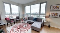 This contemporary high-rise residence offers a bright, open layout defined by large floor-to-ceiling windows that frame sweeping panoramic views of the surrounding cityscape. The living area is a standout feature, with expansive corner windows flooding the space with natural light and providing unobstructed vistas of the urban skyline. Light hardwood flooring runs throughout the main living areas, complementing the clean, modern aesthetic.

The kitchen is thoughtfully designed with sleek white cabinetry extending to the ceiling for ample storage, granite countertops, and a full suite of stainless steel appliances including a refrigerator, range, microwave, and dishwasher. A hallway connects the living areas to the private quarters of the residence.

Two well-proportioned bedrooms offer comfortable retreats, each with plush carpeting. One bedroom features a closet and a large window with city views, while the other provides a quiet, restful ambiance with stylish wall accents.

The residence includes two bathrooms: one outfitted with a tiled walk-in shower and granite vanity, and the other featuring a full soaking tub with tiled surround, tile flooring, and a granite-topped vanity. Together, these spaces deliver a polished, move-in-ready home with modern finishes throughout.