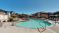 Located at 5959 S Cougar Ln in Kearns, Utah, this single-floor community clubhouse offers a well-appointed gathering space designed with a contemporary aesthetic. The interior opens through a hallway featuring striking geometric hardwood-patterned flooring, leading into a bright, open living room with soaring ceilings accented by warm wood paneling. The living room is thoughtfully furnished with a mix of modern sofas, a natural wood coffee table, and curated abstract artwork, creating a welcoming and stylish atmosphere. Floor-to-ceiling glass partitions divide the interior spaces, allowing natural light to flow freely throughout. A dedicated game room, visible through the glass walls, showcases a dramatic floor-to-ceiling ocean mural and a full-size pool table, adding a vibrant, recreational dimension to the space. High-top bistro tables and bar seating provide additional gathering spots within the clubhouse. Outside, the expansive amenity deck features a large resort-style swimming pool surrounded by lounge chairs and teal umbrellas. A covered pergola with ceiling fans offers shaded seating, while a built-in stone outdoor kitchen with a grill provides space for al fresco entertaining. Landscaped grounds with ornamental grasses and open sun-drenched patio areas complete this impressive community amenity space.