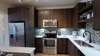 The kitchen is designed with a contemporary aesthetic, showcasing rich dark wood-grain cabinetry that extends from floor to ceiling, maximizing storage throughout the space. The upper and lower cabinets are finished with sleek bar-pull hardware, contributing to the clean, modern look. Stainless steel appliances, including an electric range, over-the-range microwave, and refrigerator, provide a polished, professional feel. The countertops feature a crisp white finish that contrasts effectively against the darker cabinetry, enhancing the visual balance of the kitchen. A standout design element is the mosaic tile backsplash, which incorporates shades of blue, grey, and white in a linear brick pattern, adding texture and a subtle pop of color. Light wood-tone flooring runs through the space, softening the overall palette and adding warmth. Recessed track lighting in the ceiling provides focused illumination across the workspace. The kitchen is efficiently laid out in an L-shaped configuration, making practical use of the available space while maintaining an open and accessible flow.