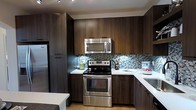 The kitchen is designed with a contemporary aesthetic, showcasing rich dark wood-grain cabinetry that extends from floor to ceiling, maximizing storage throughout the space. The upper and lower cabinets are finished with sleek bar-pull hardware, contributing to the clean, modern look. Stainless steel appliances, including an electric range, over-the-range microwave, and refrigerator, provide a polished, professional feel. The countertops feature a crisp white finish that contrasts effectively against the darker cabinetry, enhancing the visual balance of the kitchen. A standout design element is the mosaic tile backsplash, which incorporates shades of blue, grey, and white in a linear brick pattern, adding texture and a subtle pop of color. Light wood-tone flooring runs through the space, softening the overall palette and adding warmth. Recessed track lighting in the ceiling provides focused illumination across the workspace. The kitchen is efficiently laid out in an L-shaped configuration, making practical use of the available space while maintaining an open and accessible flow.
