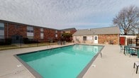 This single-floor space is part of a brick residential complex that offers access to a notable outdoor amenity area centered around a generously sized swimming pool. The pool area is finished with a wide concrete deck, providing ample space for lounging and socializing. Multiple sets of outdoor table-and-chair arrangements are positioned poolside, making the area well-suited for outdoor gatherings. The pool is enclosed by a black iron fence, adding both security and a defined boundary to the space, with open views of surrounding trees and the broader neighborhood beyond.

A dedicated pool house or utility structure is situated adjacent to the pool deck, constructed in matching brick with a gabled roof and white-trimmed windows, offering a cohesive architectural aesthetic throughout the outdoor space. Entry to the pool area is provided through a white-framed glass-paned door, lending a classic touch to the transition between interior and exterior.

Inside, the interior hallway features neutral white walls, carpeted flooring, and a series of white doors leading to individual rooms, presenting a clean and functional layout. Natural light filters into the interior spaces, casting soft shadows across the walls. The overall ambiance of the space is practical and straightforward, with key amenities conveniently accessible to residents.