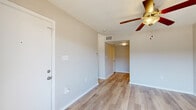 This single-floor unit offers a clean, functional layout with a cohesive design throughout. The main living area is characterized by light wood-look flooring that extends across the space, neutral light gray walls, and a ceiling fan with a built-in light fixture that provides both airflow and ambient lighting. A sliding glass door fills the room with natural light and opens to a private balcony, offering a pleasant outdoor extension of the living space.

The entry area includes a standard door with a light switch panel and HVAC vent, keeping climate control accessible. A hallway connects the living area to the remaining rooms, maintaining a straightforward and efficient floor plan.

The kitchen is equipped with white cabinetry, granite-style countertops, and black appliances including a refrigerator, electric range with overhead microwave/vent hood, and dishwasher. The light flooring continues into this space, maintaining visual consistency.

The bathroom features wood-look tile flooring, a full tub and shower combination with white square tile surround, brushed nickel fixtures and towel bar, and a toilet, offering a clean and practical design. Overall, the unit presents a well-maintained, move-in ready space with consistent finishes throughout.