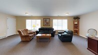 This single-story space is designed to serve both administrative and communal functions, offering a practical and welcoming environment throughout. The main lounge area is generously proportioned, featuring wall-to-wall carpet flooring, warm beige walls, and multiple large windows with vertical blinds that allow ample natural light to fill the room. Seating includes a full leather sofa, a leather loveseat, and an upholstered armchair arranged around a wooden coffee table, creating a comfortable gathering area. A corner bookshelf unit provides additional storage and display space.

An adjoining open area extends the usable footprint considerably, featuring a ceiling fan, additional flush-mount lighting, and framed artwork on the walls. This flexible zone accommodates a workstation with a desk and seating, a small children's table and chair set, and wall-mounted informational panels, suggesting a multi-use community function.

A dedicated office area is anchored by a substantial dark wood executive desk paired with a leather chair, a decorative potted tree, and direct access to an exterior entry door. A kitchenette with cabinetry is also visible nearby. Overall, the space presents a functional, organized, and accessible environment suited for office or community use.