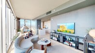 Located in the heart of Downtown Los Angeles at 100 S Grand Ave, this high-rise residence offers a sophisticated single-floor layout defined by floor-to-ceiling glass windows that frame breathtaking panoramic views of the city skyline and the San Gabriel Mountains beyond. Natural light floods every corner of the open-plan living and dining area, where a curved sofa, marble-top coffee table, and an artistic pendant light create a refined yet comfortable atmosphere.

The kitchen features sleek, handle-free cabinetry in a light wood tone, white quartz countertops, and professional-grade appliances, including a gas range. A peninsula connects the kitchen seamlessly to the main living space. The bedroom includes a textured stone accent wall, floor-to-ceiling windows, and ample natural light, while a secondary room serves as a versatile lounge or sitting area with equally impressive city views.

The bathrooms are finished to a high standard: one features full marble-slab walls within a spacious glass-enclosed shower, while another offers a soaking tub with stone tile surround. Light wood flooring flows throughout, and a crisp, softly toned hallway connects each room with cohesion and calm.