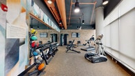 Located at 411 W Eighth St in Bloomington, Indiana, this exercise room is a well-appointed fitness center that combines industrial-chic aesthetics with a comprehensive array of workout equipment. The space is defined by its striking design elements: a dark ceiling with exposed copper ductwork and pendant lighting creates a bold, urban atmosphere, while a vibrant geometric mural adds a dynamic splash of color along one wall. Textured brick accent walls and large concrete structural columns contribute to the room's distinctive character.

Floor-to-ceiling windows line one side of the fitness center, flooding the space with natural light and offering views of the surrounding greenery. Roller shades provide adjustable light control. Durable speckled rubber flooring covers the entire area, offering both safety and practicality for exercise.

The equipment selection is thorough and well-organized. Cardio options include True-brand treadmills and elliptical machines, as well as a rowing machine. A dedicated strength zone features a full dumbbell rack with kettlebells, a multi-function cable machine, and adjustable weight benches. Medicine balls of varying weights are neatly stored on a rack near a built-in wooden bench with cubby storage. Wall-mounted televisions and ceiling fans complete the amenity-rich environment, making this a fully functional and visually engaging fitness facility.