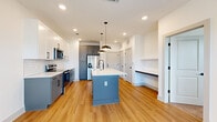 This contemporary single-floor residence showcases a thoughtfully designed open-concept layout where the kitchen and living area flow seamlessly together. The kitchen is a standout feature, anchored by a generously sized center island with a white quartz countertop, an undermount sink, and a matte black pull-down faucet. Two-tone cabinetry — white uppers paired with slate-blue lowers — lines the walls alongside a classic white subway tile backsplash. Stainless steel appliances, including a French door refrigerator, range, microwave, and dishwasher, complete the space. Stylish matte black pendant lights hang above the island, complementing the modern hardware throughout. The living area extends naturally from the kitchen, featuring warm-toned hardwood-style flooring, a ceiling fan, and a glass door that opens to a private balcony, filling the space with abundant natural light and outdoor views. Bedrooms offer plush carpeting and well-sized windows that bring in ample daylight. The bathroom is finished with a white shaker-style vanity, white quartz countertop, matte black fixtures, a large mirror, and access to a walk-in closet with organizational shelving. Crisp white walls and recessed lighting throughout the unit create a clean, bright, and welcoming ambiance.