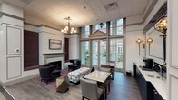 The lounge at 480 Bedford Rd in Chappaqua, New York, is a thoughtfully designed communal space that blends classical architectural details with modern, upscale furnishings. The single-floor layout is anchored by soaring clerestory windows topped with a decorative pediment, flooding the interior with natural light and offering unobstructed views of the surrounding courtyard. A set of French doors provides direct access to the outdoor patio, seamlessly connecting the interior to the exterior environment.

The seating area is defined by rich hardwood-style flooring and a patterned area rug, upon which a curated arrangement of leather armchairs, a tufted bench, and geometric side tables are arranged. A decorative fireplace with an ornate tile surround and a mantel serves as a focal point on one wall, with a framed artwork displayed above it. A multi-arm chandelier overhead adds a refined, traditional touch to the space.

Adjacent to the lounge seating, a dining area features marble-topped tables paired with upholstered chairs, offering a more structured setting for casual gatherings. A bar-style counter with dark subway tile backsplash and a wall-mounted mirror provides a beverage station. Large glass doors on the opposite side of the lounge reveal direct sightlines into a fitness center, further enhancing the sense of openness and connectivity throughout the space.