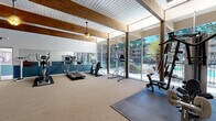 This fitness and recreation center offers a spacious, open layout designed to accommodate a full range of exercise and leisure activities. The architecture is defined by striking exposed wooden beams that span the length of the vaulted ceiling, complemented by a white tongue-and-groove ceiling finish and warm pendant lighting that creates an inviting ambiance throughout the space.

One of the room's most prominent features is the floor-to-ceiling glass wall along one side, which floods the interior with abundant natural light and provides expansive views of the outdoor pool area and surrounding greenery. The neutral carpet and rubber mat flooring zones provide practical surfaces for both cardio and strength training.

The fitness area is well-appointed with a treadmill, elliptical trainer, stationary bike, cable machine, weight bench, and a full dumbbell rack with a range of free weights. A large wall mirror enhances the sense of openness while supporting proper form during workouts. Instructional fitness posters and a stability ball round out the training amenities.

Beyond fitness, the space includes a foosball table and an upright piano, adding a recreational and social dimension. Two-tone blue and white walls lend a clean, polished look, while French doors provide an additional access point. This multifunctional facility is thoughtfully designed to serve both active and leisure needs.