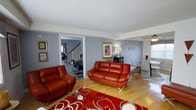 This two-floor home offers a thoughtfully designed interior that balances bold style with functional living. The main level centers around an open living area finished with warm hardwood floors that extend throughout the space, creating a cohesive and inviting atmosphere. The living room makes a strong visual statement with rich cognac leather sofas, a vibrant red floral area rug, and carefully curated wall art — including a prominent gold-framed piece that anchors the room's aesthetic. Soft gray walls provide a neutral backdrop that complements the warm tones of the furnishings. A ceiling fan overhead adds both practicality and a subtle architectural detail. The layout flows openly into a dining area, where a modern white dining set and sleek chairs offer an elegant contrast to the warmer tones of the living space. Framed artwork and a classic table lamp add refined finishing touches throughout. An open doorway reveals a staircase leading to the second floor, as well as a glimpse of an additional dining or multi-purpose area beyond. The interconnected layout of the main floor promotes a natural and easy flow between spaces, making this home well-suited for both everyday living and entertaining.
