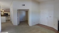 This single-story space presents a straightforward, functional layout with a clean and neutral aesthetic throughout. The main room features wall-to-wall carpet in a warm beige tone, contributing to a soft, understated ambiance. The walls are finished in a light, off-white color that reflects available light and creates a sense of openness. Notably, the ceilings rise higher than standard, adding vertical volume to the space and enhancing the overall sense of airiness.

An open doorway leads directly into an adjoining room, also carpeted and similarly finished, providing a seamless flow between the two areas. A second door, featuring a classic panel design, opens to a separate area where tile flooring is visible, likely indicating a transitional space such as an entryway or utility area. An HVAC vent is positioned near the upper portion of the wall, suggesting a central climate control system is in place.

The space is currently vacant, allowing for a clear assessment of its structural features. The overall condition appears well-maintained, with clean walls and flooring throughout. The neutral palette and simple design provide a versatile foundation suitable for a variety of uses and interior arrangements.