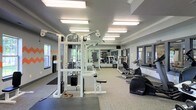 This fitness center offers a spacious, single-floor layout designed to accommodate a comprehensive workout experience. The room is finished in a neutral gray color palette, lending a clean and modern aesthetic throughout. Overhead fluorescent lighting ensures the space is consistently well-lit, complementing the natural light that filters in through multiple windows along the walls.

The strength training area is anchored by a large multi-functional cable machine, providing users with a wide range of resistance exercise options. Additional weight and resistance equipment is arranged throughout the space, allowing multiple users to train simultaneously without congestion. A wall of mirrors runs along one side of the room, enhancing the sense of depth while allowing users to monitor their form.

The cardio section features equipment such as a rowing machine and other aerobic machines positioned toward the perimeter. Rubber floor mats are placed in key areas to provide traction and protect flooring during workouts. The layout is thoughtfully organized to maintain clear pathways between equipment zones, supporting both safety and ease of movement throughout the facility.