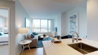 This single-floor residence is designed with a contemporary aesthetic, combining clean lines, neutral tones, and high-quality finishes throughout. The open-concept living area is anchored by floor-to-ceiling windows that frame sweeping waterfront and city skyline views, flooding the space with natural light. Soft blue-grey walls complement light hardwood flooring, creating a calm, cohesive atmosphere. The living area accommodates a comfortable sectional sofa, a round coffee table, and a teal area rug that adds a pop of color.

The kitchen is efficiently laid out with white shaker-style cabinetry extending to the ceiling for ample storage, stainless steel appliances including a professional-grade gas range and refrigerator, and a marble-toned countertop that extends into a breakfast bar. The transition between kitchen and living space feels seamless, supporting an open and social floor plan.

The bedroom is a tranquil retreat, featuring plush carpeting, a textured accent wall, and large windows offering elevated city and water views. A full-length mirror and greenery add depth and character to the space. The bathroom is finished in crisp white, with a dual-sink vanity topped in marble-style stone, tall cabinetry for storage, and illuminated mirrors that enhance the bright, clean aesthetic.